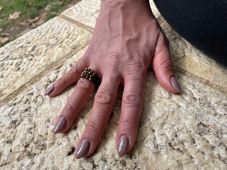 K&ouml;nnte beinhalten: Eine Hand tr&auml;gt einen goldenen Ring mit mehreren Reihen kleiner, dunkelbrauner Steine. Die N&auml;gel sind in einem hellen Braunton lackiert.