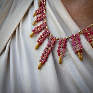 May include: A pink and gold beaded necklace with a geometric pattern. The necklace is made of small, square beads that are arranged in a repeating pattern. The beads are strung on a thin, gold chain.