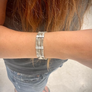 May include: Silver bracelet with multiple strands, featuring a mix of textured and smooth links. The bracelet is worn on a person's arm, showcasing its intricate design and metallic sheen. The bracelet appears to be made of silver.