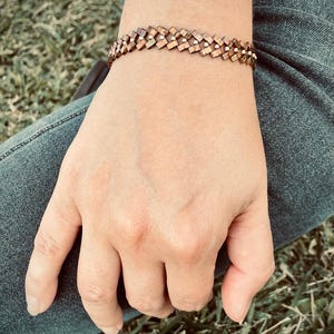 May include: A copper bracelet with a woven design. The bracelet is made of small, square-shaped pieces of copper that are interlocked to create a geometric pattern.