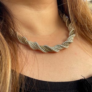 Puede incluir: Un collar de cadena trenzado de plata y oro. El collar está hecho de pequeñas cuentas y lo lleva una persona.