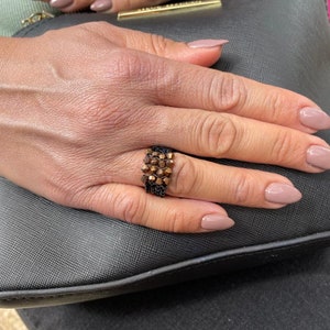 K&ouml;nnte beinhalten: Nahaufnahme einer Hand, die einen Ring mit braunem und schwarzem Perlenmuster tr&auml;gt. Der Ring befindet sich am Ringfinger der rechten Hand. Die Hand ruht auf einer schwarzen, strukturierten Oberfl&auml;che.