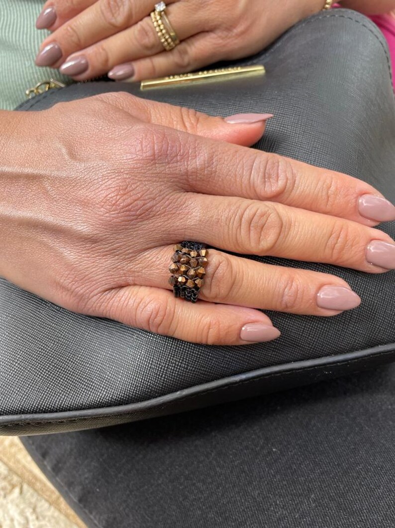 K&ouml;nnte beinhalten: Nahaufnahme einer Hand, die einen Ring mit braunem und schwarzem Perlenmuster tr&auml;gt. Der Ring befindet sich am Ringfinger der rechten Hand. Die Hand ruht auf einer schwarzen, strukturierten Oberfl&auml;che.