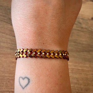 May include: A brown and gold bracelet with a geometric pattern. The bracelet is made of small, square beads that are arranged in a repeating pattern. The beads are interspersed with small, round gold beads.