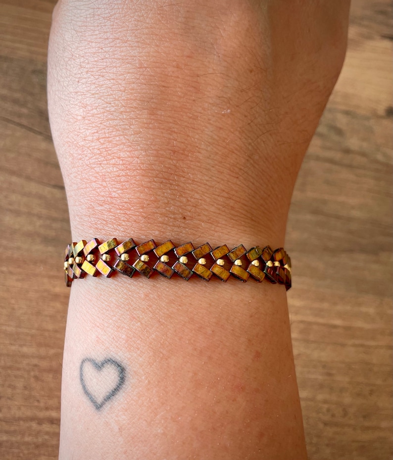 May include: A brown and gold bracelet with a geometric pattern. The bracelet is made of small, square beads that are arranged in a repeating pattern. The beads are interspersed with small, round gold beads.