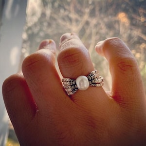 May include: A silver ring with a large white pearl in the center. The ring is made of multiple bands of silver beads.