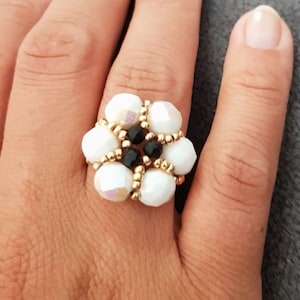 Könnte beinhalten: Ein weißer, blumenförmiger Ring mit schwarzen Perlen in der Mitte. Der Ring ist aus goldfarbenem Metall und wird an einem Finger getragen.