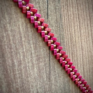 May include: A close-up of a pink and gold beaded necklace. The beads are arranged in a chevron pattern.