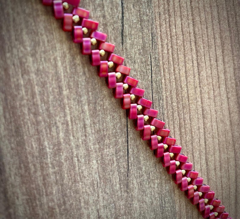 May include: A close-up of a pink and gold beaded necklace. The beads are arranged in a chevron pattern.