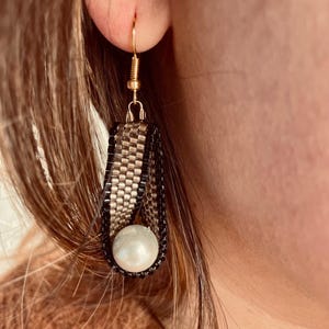 May include: A pair of gold and black beaded earrings with a white pearl accent. The earrings are dangling and have a simple gold hook fastening.
