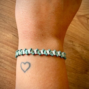 May include: A turquoise and white beaded bracelet on a person's wrist. The bracelet is made of small, square beads.