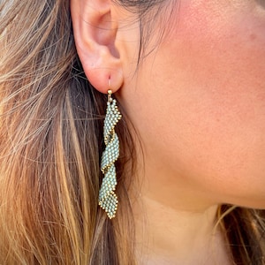 May include: A pair of long, dangling earrings with a twisted design. The earrings are made of small, light blue beads and gold wire.