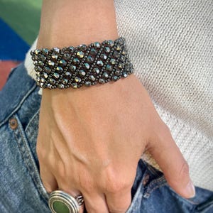 May include: A wide cuff bracelet composed of multiple rows of faceted, metallic dark gray beads arranged in a diagonal pattern. A ring featuring a circular green stone set in a silver-toned metal frame is also visible on the finger.