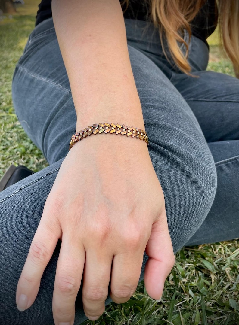 May include: A copper-toned bracelet with a woven design. The bracelet is made of small, square-shaped links.