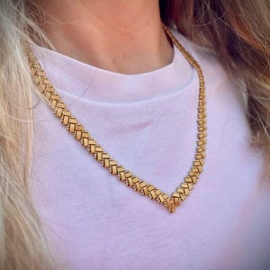 May include: A gold-colored necklace with a woven design. The necklace is worn on a person wearing a white t-shirt. The necklace has a V-shaped pendant and is made of small, rectangular links.