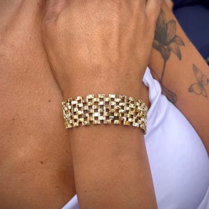 May include: A wide gold-toned bracelet featuring a repeating, interlocking rectangular grid pattern. The metal surface has a textured, reflective finish with alternating light and dark metallic highlights across the woven-style link structure.