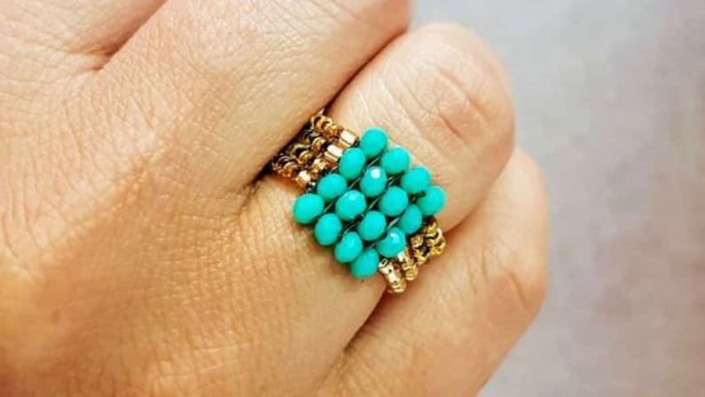 K&ouml;nnte beinhalten: Ein t&uuml;rkisfarbener Perlenring mit goldenen Akzenten. Der Ring ist aus kleinen, runden Perlen gefertigt und wird an einem Finger getragen.