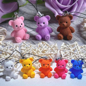 Könnte beinhalten: Zehn gehäkelte Teddybär-Schlüsselanhänger in verschiedenen Farben, darunter rosa, lila, braun, weiß, gelb, orange, rot und blau. Die Schlüsselanhänger hängen an einer weißen Oberfläche.