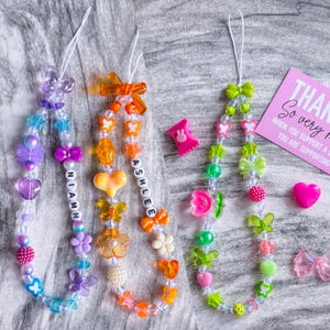 Puede incluir: Encantos de teléfono con cuentas de colores con varias formas, incluyendo corazones, mariposas y flores. Un encanto deletrea "Niamh" y otro "Ashlee" con cuentas de letras. También se ven encantos adicionales y un corazón rosa.