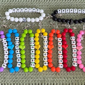 Può includere: Braccialetti colorati con perline che compongono la parola "BREATHE" con perline di lettere bianche. I braccialetti sono in vari colori, tra cui bianco, nero, viola, blu, verde, giallo, arancione e rosa. Alcuni braccialetti hanno perline a forma di cuore.