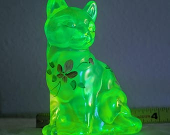 May include: A glowing green cat figurine with floral accents. The cat is in a seated position, with its ears perked up. The figurine is approximately 4 inches tall.