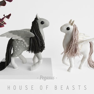 Peut inclure: Deux figurines Pegasus faites à la main en feutre. L'une est grise avec une crinière et une queue noires, l'autre est blanche avec une crinière et une queue marron clair. Les deux ont des ailes et sont debout. Le texte "Pegasus" et "HOUSE OF BEASTS" est en dessous.