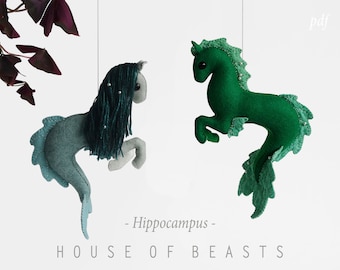 May include: Two felt seahorse ornaments hanging from strings. The seahorses are green and blue with a white belly and a green and blue tail. The text "Hippocampus" and "HOUSE OF BEASTS" is printed below the seahorses.