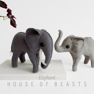 May include: Two gray felt elephant plushies, one larger than the other, with stitched details and black button eyes. The larger elephant has white felt tusks. The plushies are on a white surface with the text "HOUSE OF BEASTS - Elephant -" below them.