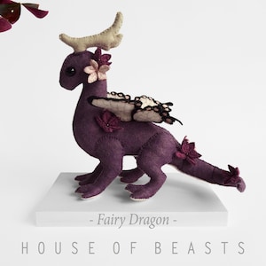 May include: Purple felt fairy dragon with butterfly wings and flower accents. The dragon is standing on a white platform with the text "Fairy Dragon" and "HOUSE OF BEASTS" printed below.