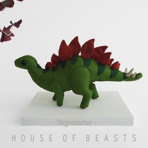 May include: A green felt Stegosaurus plush toy with red felt plates on its back. The toy is standing on a white platform with the text "HOUSE OF BEASTS - Stegosaurus -" printed on it.