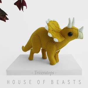 May include: A yellow felt Triceratops plush toy with white felt accents. The toy is standing on a white surface with the text "HOUSE OF BEASTS - Triceratops -" printed below it.