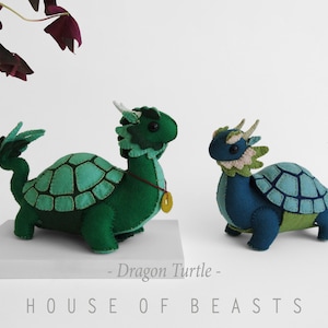 May include: Two felt dragon turtle plushies, one green and one blue, with a green and blue shell pattern. The plushies are sitting on a white surface with the text "Dragon Turtle" and "HOUSE OF BEASTS" below them.