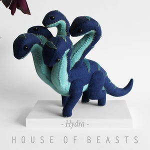 Peut inclure: Une peluche hydre bleue à quatre têtes, avec un ventre et des détails turquoise. La créature faite à la main est posée sur une plateforme blanche, avec le texte "- Hydra - HOUSE OF BEASTS" en dessous. Une créature mythique.