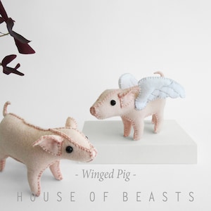May include: Two felt pig plushies, one with white wings. The winged pig is standing on a white shelf. The text "Winged Pig" is below the shelf. The text "HOUSE OF BEASTS" is below the text "Winged Pig".