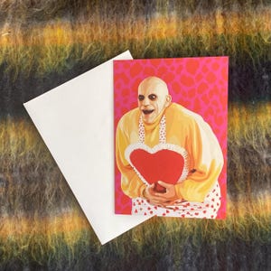 May include: A Valentine's Day card featuring a cartoon character holding a red heart-shaped cushion. The character is wearing a yellow shirt with a white apron and a white collar. The background is pink with white hearts.