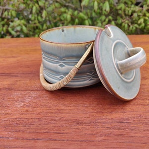 Puede incluir: Un recipiente de cerámica con tapa, con un esmalte azul verdoso y un asa tejida. El recipiente tiene un patrón decorativo alrededor del centro. La tapa tiene un asa de bucle. El borde del recipiente y la tapa son marrones.