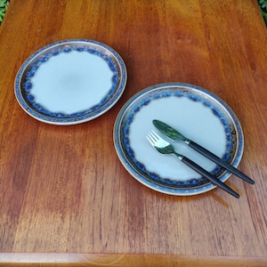 May include: Two light blue ceramic plates with a dark blue and gold rim. One plate has a fork and knife with dark brown handles. The plates are on a wooden surface.
