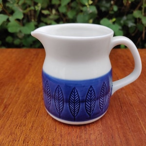 Puede incluir: Una jarra de crema de cerámica blanca con un patrón de hojas azul y blanco. La jarra tiene un cuerpo redondeado y un asa.