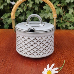 Puede incluir: Un azucarero de cerámica blanca con un diseño de rejilla marrón y un asa de madera. El tazón está sobre una superficie de madera.