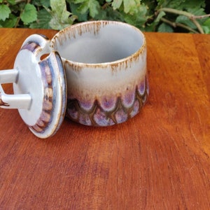 Puede incluir: Un azucarero de cerámica con tapa y asa. El bol está decorado con un esmalte marrón, blanco y morado. La tapa tiene un pequeño agujero para verter.