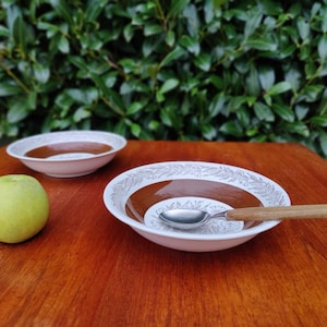 May include: Two white ceramic bowls with brown and white floral patterns, one bowl is empty and the other has a silver spoon inside. A green apple sits on a wooden table.