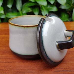 Puede incluir: Un azucarero de cerámica blanca con un borde marrón y una tapa a juego. La tapa tiene una pequeña asa y un pico para verter.