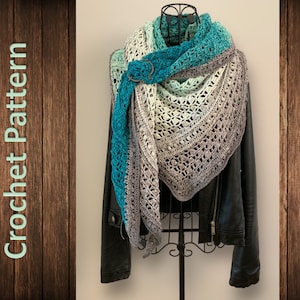 Peut inclure: Un châle en crochet dans des tons de turquoise, vert et gris avec une boucle décorative. Le châle est drapé sur une veste en cuir noir sur un mannequin.