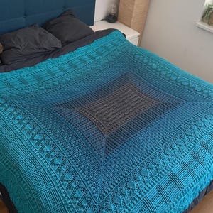 Könnte beinhalten: Eine türkisfarbene, blaue und graue gemusterte Decke bedeckt ein Bett. Die Decke hat ein geometrisches Design mit einem zentralen Quadrat und aufwendigen Bordüren. Der Farbverlauf geht von Türkis über Blau zu Grau. Ein Teddybär sitzt auf dem Bett.