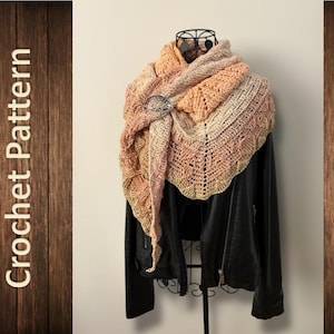 May include: A crocheted shawl in peach, cream, and tan, fastened with a decorative brooch. The shawl is draped over a black leather jacket. The words "Crochet Pattern" are vertically displayed on a wooden background.