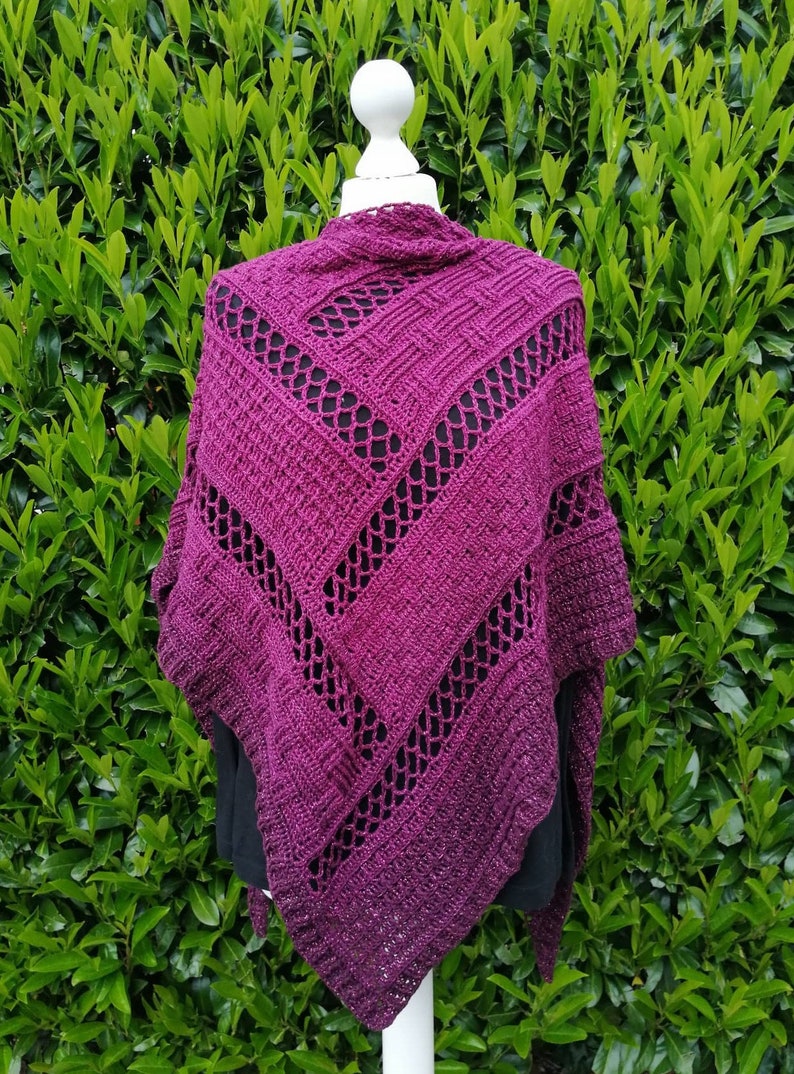 Puede incluir: Un chal de ganchillo morado con un patr&oacute;n de encaje. El chal est&aacute; drapeado sobre un maniqu&iacute; blanco y se muestra sobre un fondo de follaje verde.