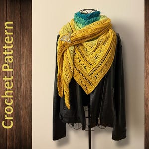 Op de afbeelding: Een gele, groene en blauwe gehaakte sjaal met een decoratieve broche. De sjaal is over een zwart leren jack gelegd.