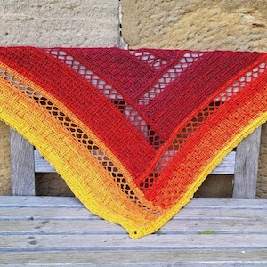 Puede incluir: Un chal de ganchillo triangular en tonos de amarillo, naranja y rojo. El chal est&aacute; drapeado sobre un banco de madera.