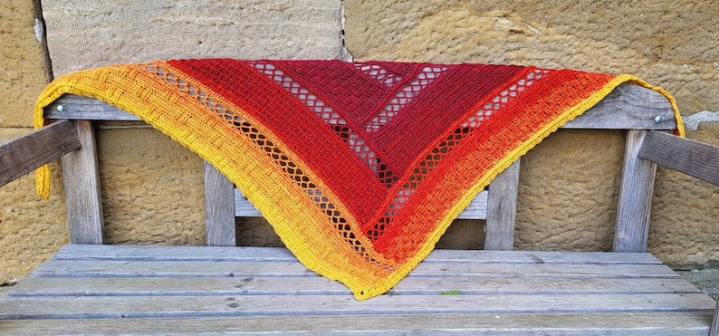 Puede incluir: Un chal de ganchillo triangular en tonos de amarillo, naranja y rojo. El chal est&aacute; drapeado sobre un banco de madera.