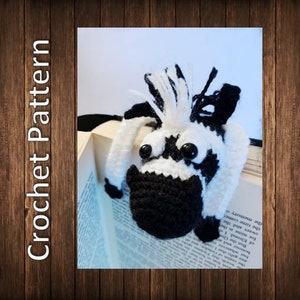 Könnte beinhalten: Ein gehäkeltes Zebra-Lesezeichen mit schwarzen und weißen Streifen und schwarzen Augen. Das Lesezeichen liegt auf einem Buch mit Text.